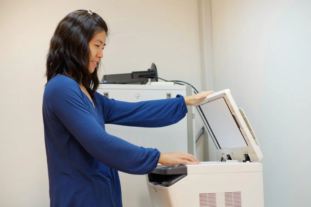 A middle aged Asian woman, working as a secretary is copying documents using a multifunction laser printer at her office.